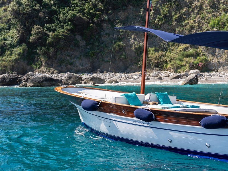 A traditional wooden gozzo with a blue awning, moored close under the rocks